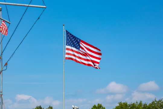American flag waving against a clear blue sky with fluffy white clouds and green trees in the background, symbolizing patriotism and freedom