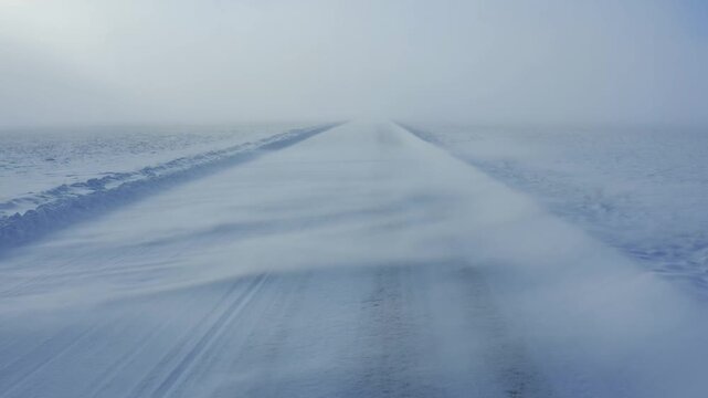 Tracking shot of snow covered road moving forward through blowing snow across frozen plain in foggy winter landscape