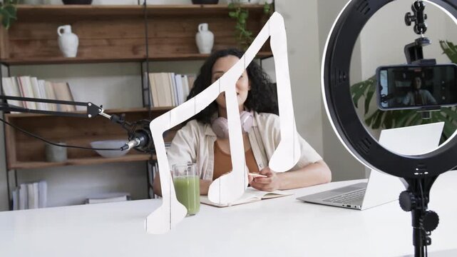 Woman recording starting phone in ringlight, speaking for podcast, music-note overlaying foreground