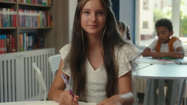 Caucasian teen girl teenager schoolgirl pupil writing notes educate study smiling looking at camera student teenager daughter kid teenaged child studying write maths exercise smile in school library