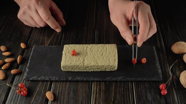 Hands skillfully use food tweezers to place red berries on a green halva dessert block placed on a slate plate, with almonds and decorative elements arranged around the culinary scene