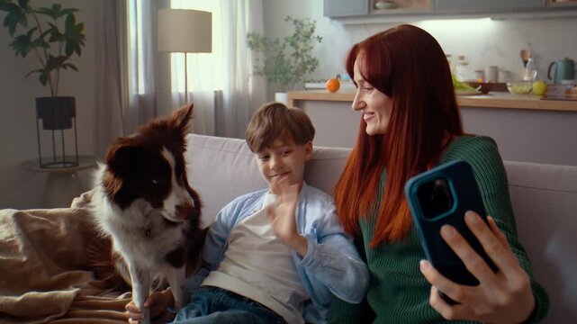 Woman waves at phone while smiling with boy and dog on sofa
