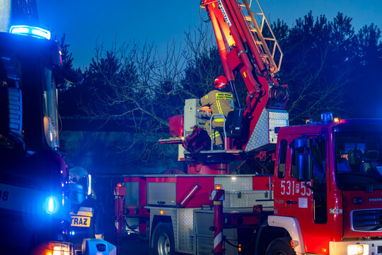 Firefighter operating a mechanical ladder on a Volvo fire engine during a night rescue operation. Professional control of a hydraulic platform with emergency blue lights flashing at dusk.
