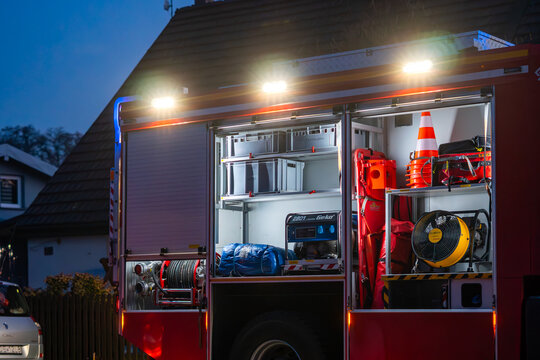 Open equipment compartment of a modern fire engine at night. Visible power generator, smoke ejector fan, traffic cones, fire hoses, and rescue tools illuminated by bright LED work lights.