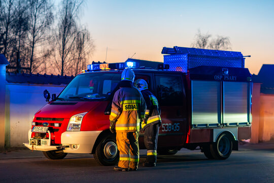 Light fire engine Ford Transit of Polish volunteer fire department at dusk with blue lights. Two firefighters in protective gear standing by the emergency vehicle during a rescue operation.