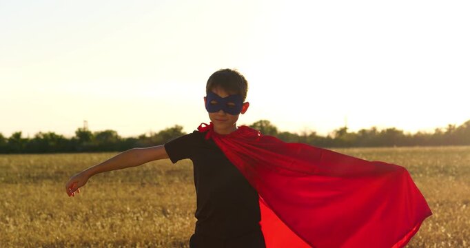 Kid playing superhero in cape and mask running outdoors in field at sunset. Happy child enjoying imaginative pretend play and freedom in nature. Kid wears mask and cape while running joyfully. Hero