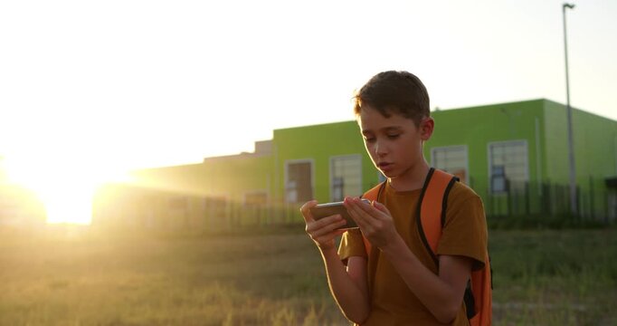 Schoolboy with backpack using smartphone after school near modern building. Concept of education, learning, and digital tools for students. Young boy uses smartphone outdoors.
