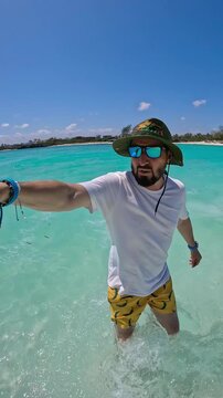 Middle-aged man wearing a sun hat and sunglasses walks through shallow turquoise ocean water while recording a selfie video in Watamu, Kenya.