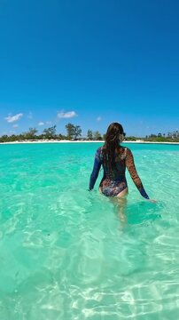 Young woman in a patterned swimsuit and sunglasses wades through clear turquoise ocean water towards a white sand beach at Watamu, Kenya.