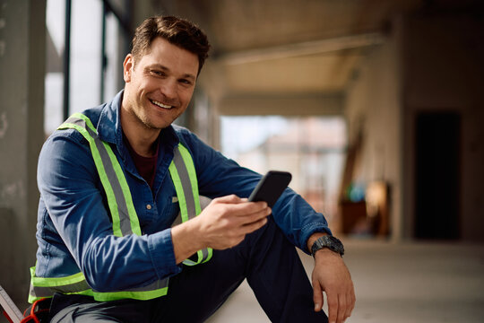 Happy worker using cell phone at construction site and looking at camera.