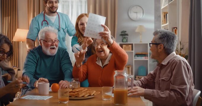 Excited elderly woman winning bingo during group game in assisted living home. Happy winner celebrating success, joy and congratulations with other seniors indoors.
