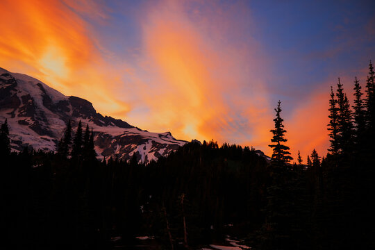 Stunning sunset over Mount Rainier, Washington. Vibrant sky with warm hues illuminating the rugged mountain peaks and surrounding forest.