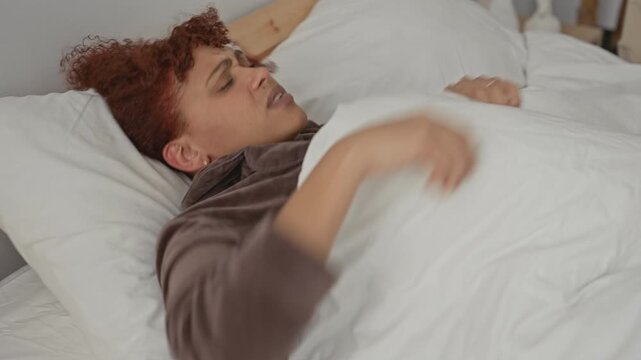 Woman clutching duvet with hand, wincing face in a small bedroom inside a building; restless discomfort.