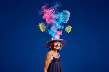 Fototapeta premium Senior woman wears iridescent hat emitting swirling pink smoke and translucent ghostly heads against solid blue background.
