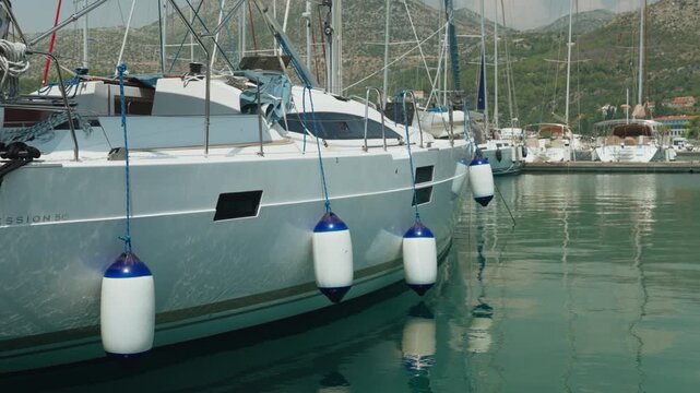 Yacht Hull With White Fenders Reflecting In Calm Marina Water, Ropes And Cleats Visible On Glossy Deck, Distant Hills And Moored Boats Create Peaceful Coastal Atmosphere