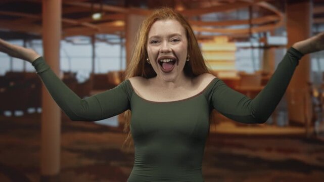 Redhead woman making hand binoculars with hands at an airport terminal, smiling and spreading arms with exposed shoulders in playful pose; joyful playfulness.