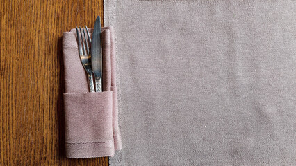 An overhead shot of a fork and knife placed in a light pink napkin, resting on a wooden table next to a patterned placemat. The composition is simple and elegant with warm lighting © Анатолий Еремин