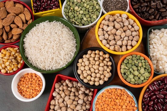 Top view of various legumes, grains, and seeds in colorful ceramic bowls including rice, lentils, chickpeas, beans, almonds, and corn, representing a healthy plant-based diet and nutrition