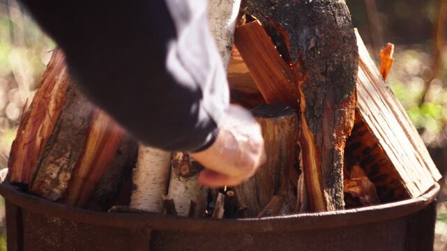 A close-up shows a man adding branches to a grill where the fire is starting to burn. The scene has natural spring light and a blurred forest background. Ideal for outdoor, camping, or BBQ content.