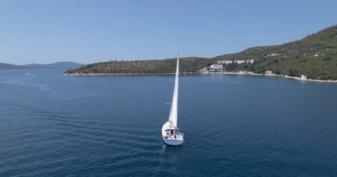 Sailboat Entering Sheltered Bay Near Rocky Headland And Low Coastal Development, Glassy Water Shows Subtle Wake And Reflections, Drone Tracks Steady Approach
