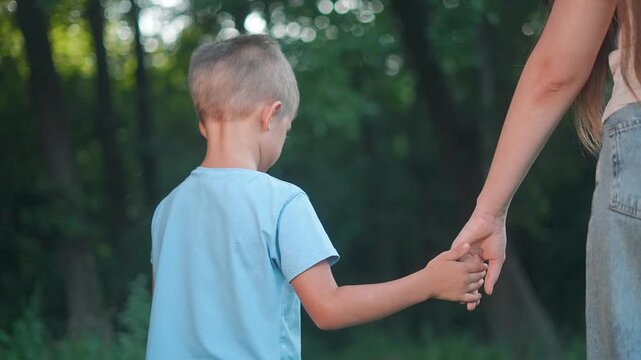Boy walks beside mother in forest. Parent hand close to boy. Outdoor walk full trust and care. Mother gives silent love. Boy moves with calm steps. Walk connects child parent through love gentle hand