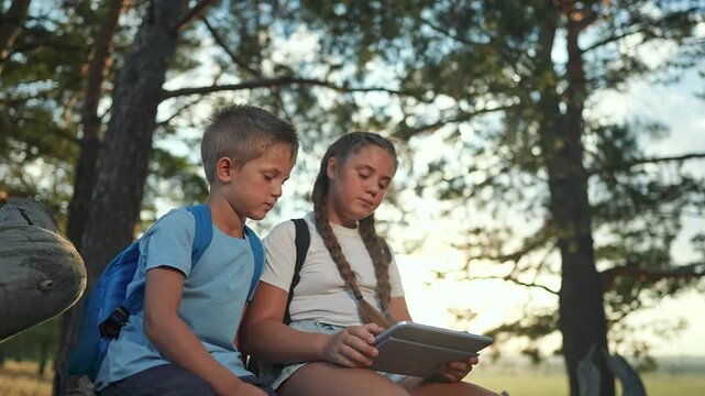 Children sit in forest using tablet together. Girl and boy explore screen sitting on log. Device in hands outdoors nature. Children tablet smile forest. Sibling use device tablet in forest sitting.