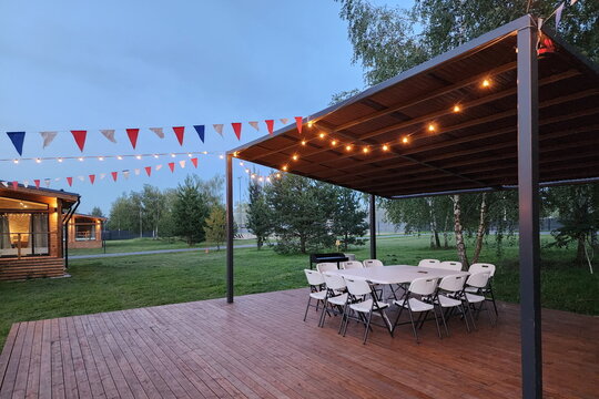 A gathering area features folding chairs set in a circle under a wooden structure. Strings of lights hang above colorful decorations. The scene takes place in an open green space at dusk