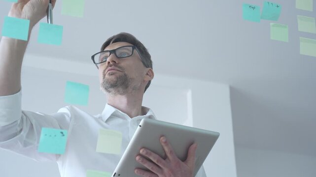 Adult businessman in glasses using a tablet computer while brainstorming and organizing work on a transparent glass board with colorful sticky notes, focusing on strategy and planning