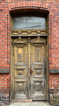 Old ornate wooden double door in a red brick wall, featuring weathered carved details, peeling paint, and an arched transom window above