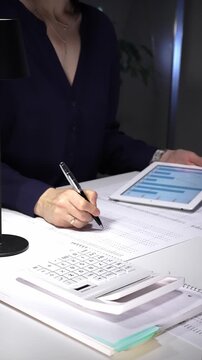 Woman working late night in office, multitasking with a tablet and writing on documents, managing heavy paperwork load. Audit and taxes in business