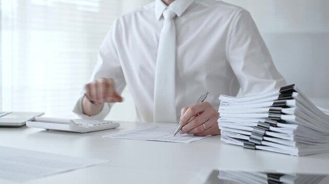 Businessman working at a desk, reviewing official documents, writing with a pen, and using a calculator, managing a stack of organized paperwork in a professional office environment. Audit and taxes