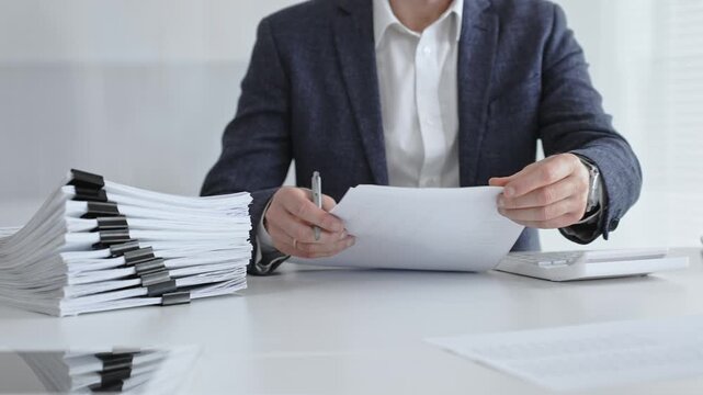 Businessman sorting through stacks of documents on a white desk beside a tall pile of bound papers, symbolizing meticulous financial record keeping and bureaucracy. Audit and taxes