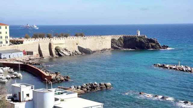 Footage of Bovio square and its lighthouse seen from the old marina, with a cruise sailing to Isle of Elba in the background, with calm sea and waves, in Piombino, Tuscany region, Italy