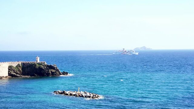 Footage of Bovio square and its lighthouse seen from the old marina, with a cruise sailing to Isle of Elba in the background, with calm sea and waves, in Piombino, Tuscany region, Italy
