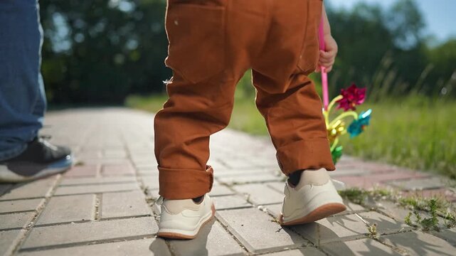 Toddler grips toy walking warm park bricks. Father follows smiling boy down sunny path sharing playful outdoor moment. Child enjoys gentle summer stroll bright pinwheel toy. Father child laugh in park
