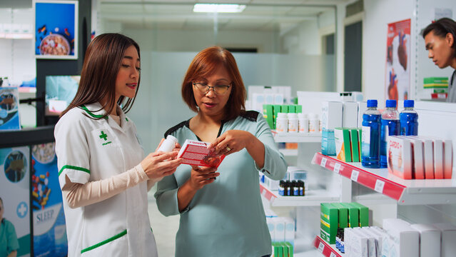 Women analyzing cardiology plls box in pharmacy shop, pharmacist giving plls and supplements to customer. Woman and drugstore worker looking at packages of healthcare medicaments.