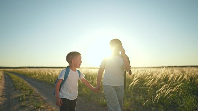 Boy and girl walking hand in hand through wide field at sunset. Child backpack shifts side to side. Girl child holds boy hand walking slow. Summer field glows. Child happy holding hand on path.