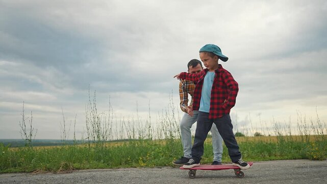 Father helps son learn to skateboard: Father supports son learning skateboarding outdoors. Kid balances on skateboard with father help. Family enjoys outdoor play while practicing skateboarding.