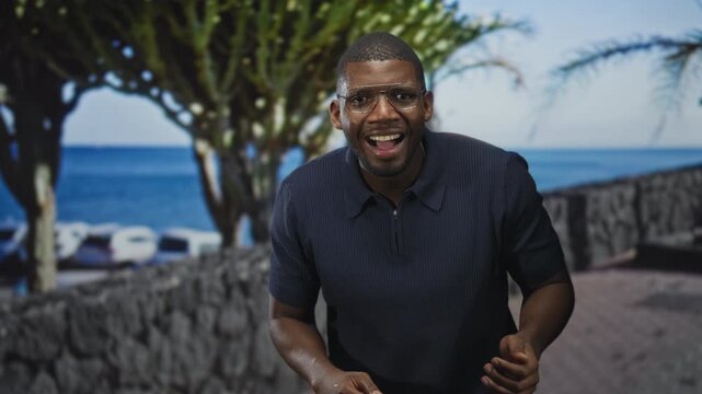 Man gesturing with hands and smiling while wearing glasses and a navy polo on a seaside street with stone wall and palm trees; joy presentation.