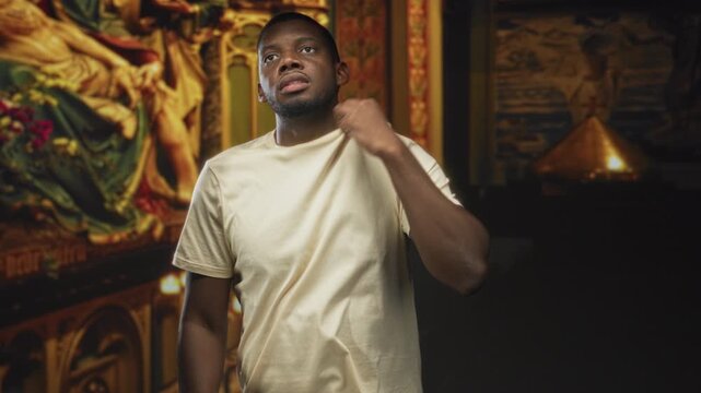 Man pulls t shirt collar toward his neck while tilting head and gazing upward beside an ornate religious mural under studio lighting; introspection.