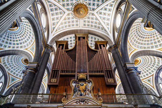 Low angle view of majestic pipe organ in choir loft of cathedral interior, framed by dark solomonic columns and ornate coffered ceilings with gold and blue details in Puebla, Mexico