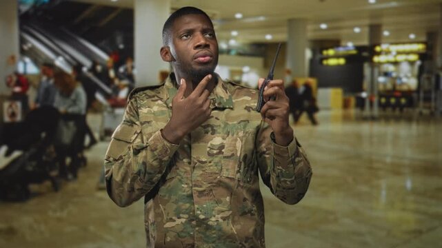 Soldier touching chin while holding radio in airport terminal, camouflage uniform visible and face focused upward; concern duty.