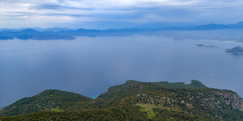 Obraz premium Sweeping aerial drone landscape looking out across a magnificent coastal bay featuring deep blue waters framed by rolling green forested mountains on a serene cloudy day