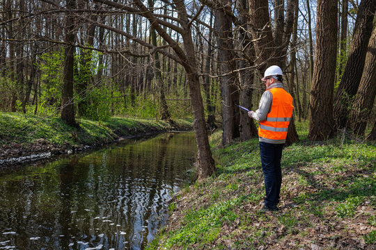 Environmental engineer or ecologist monitoring a forest river condition and water quality. Forestry specialist in safety gear standing near a river bank with a clipboard conducting ecological research