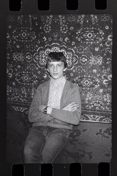 Vintage black and white portrait of young man. Retro 35mm film scan of young guy sitting in front of patterned wall carpet. Authentic USSR or Eastern Europe 80s aesthetic, nostalgia or retro fashion