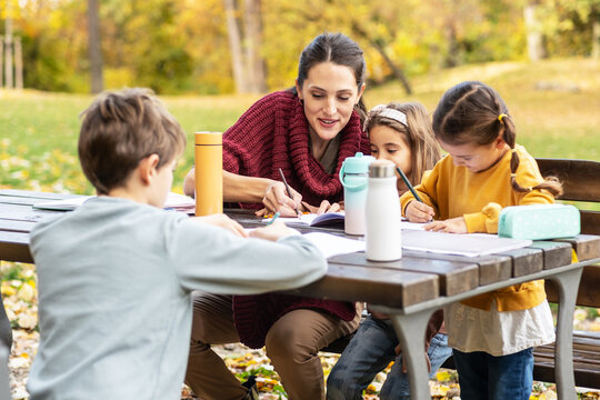 The teacher and children are now sitting together at a wooden table in the park, talking and observing nature around them. The moment is calm and focused, showing a gentle learning atmosphere outdoors