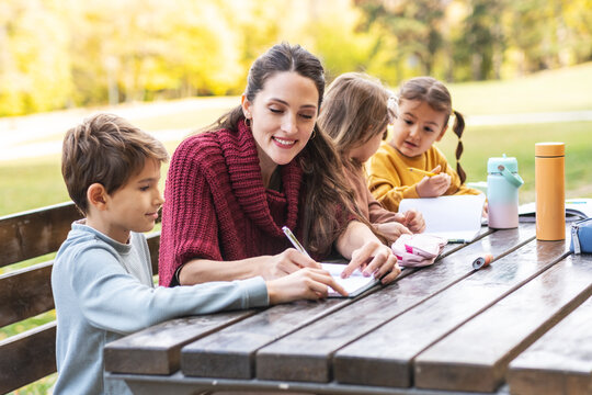 The teacher and children are now sitting together at a wooden table in the park, talking and observing nature around them. The moment is calm and focused, showing a gentle learning atmosphere outdoors