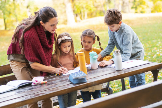 The teacher and children are now sitting together at a wooden table in the park, talking and observing nature around them. The moment is calm and focused, showing a gentle learning atmosphere outdoors