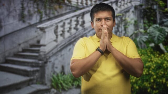Man with hands pressed together in prayer on building staircase wearing yellow polo shirt; faith reflection serenity.