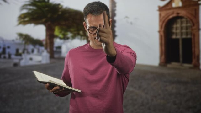 Hispanic man with glasses holding an open book points finger toward camera in a historic street; concentration.